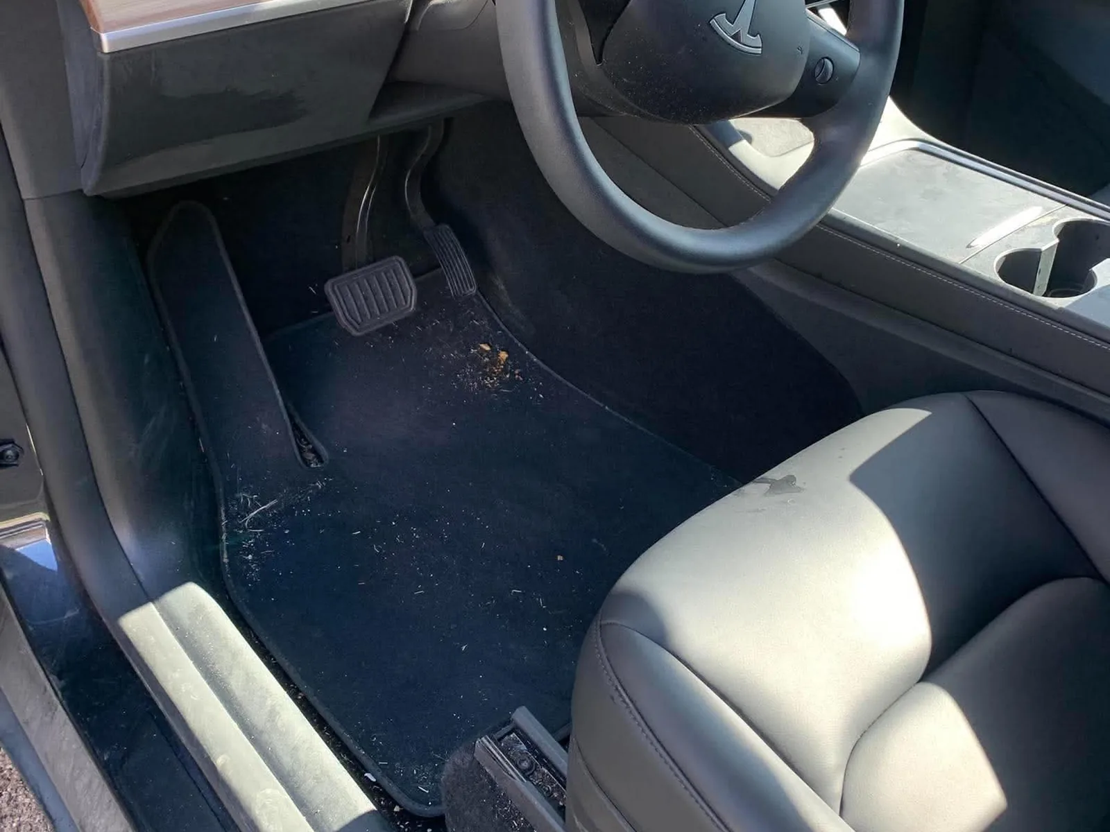 Driver footwell of an electric car before detailing, with debris scattered over the carpet and a dirty door sill
